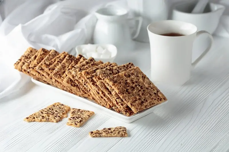 Galletas crujientes de avena y linaza apiladas junto a taza de té sobre mesa blanca