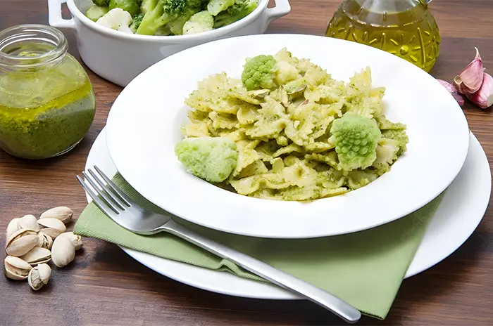 Pasta con pesto de pistachos servida en plato blanco sobre mesa de madera con aceite de oliva y ensalada
