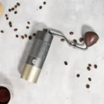 Molinillo manual de café Sweenuts Pro gris con manivela y granos de café sobre superficie clara