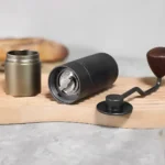Molinillo manual de café negro con ajuste de molienda y manivela desmontable sobre tabla de madera