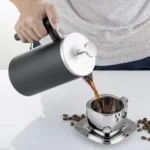 Cafetera tipo prensa francesa vertiendo café en taza de acero inoxidable sobre mesa blanca